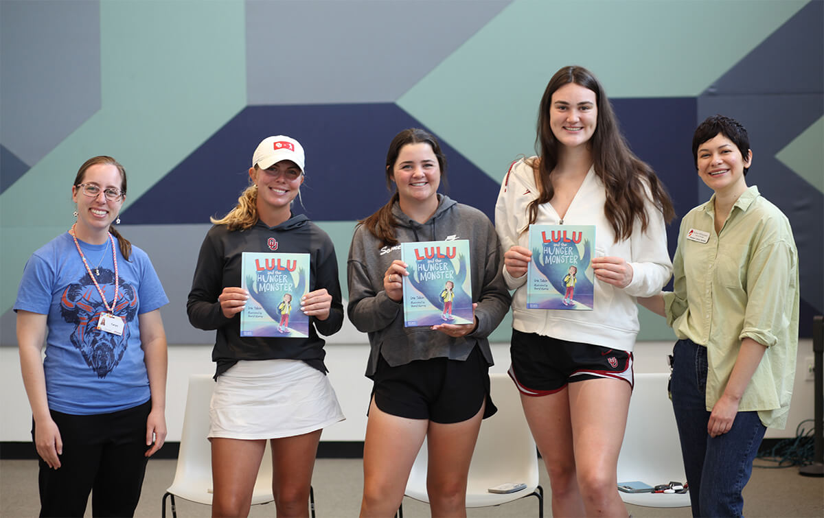 Three OU female athletes hold up the book "Lulu and the Hunger Monster". 