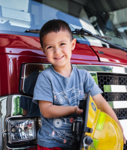 boy standing in front of a fire truck holding a firefighter's hat.